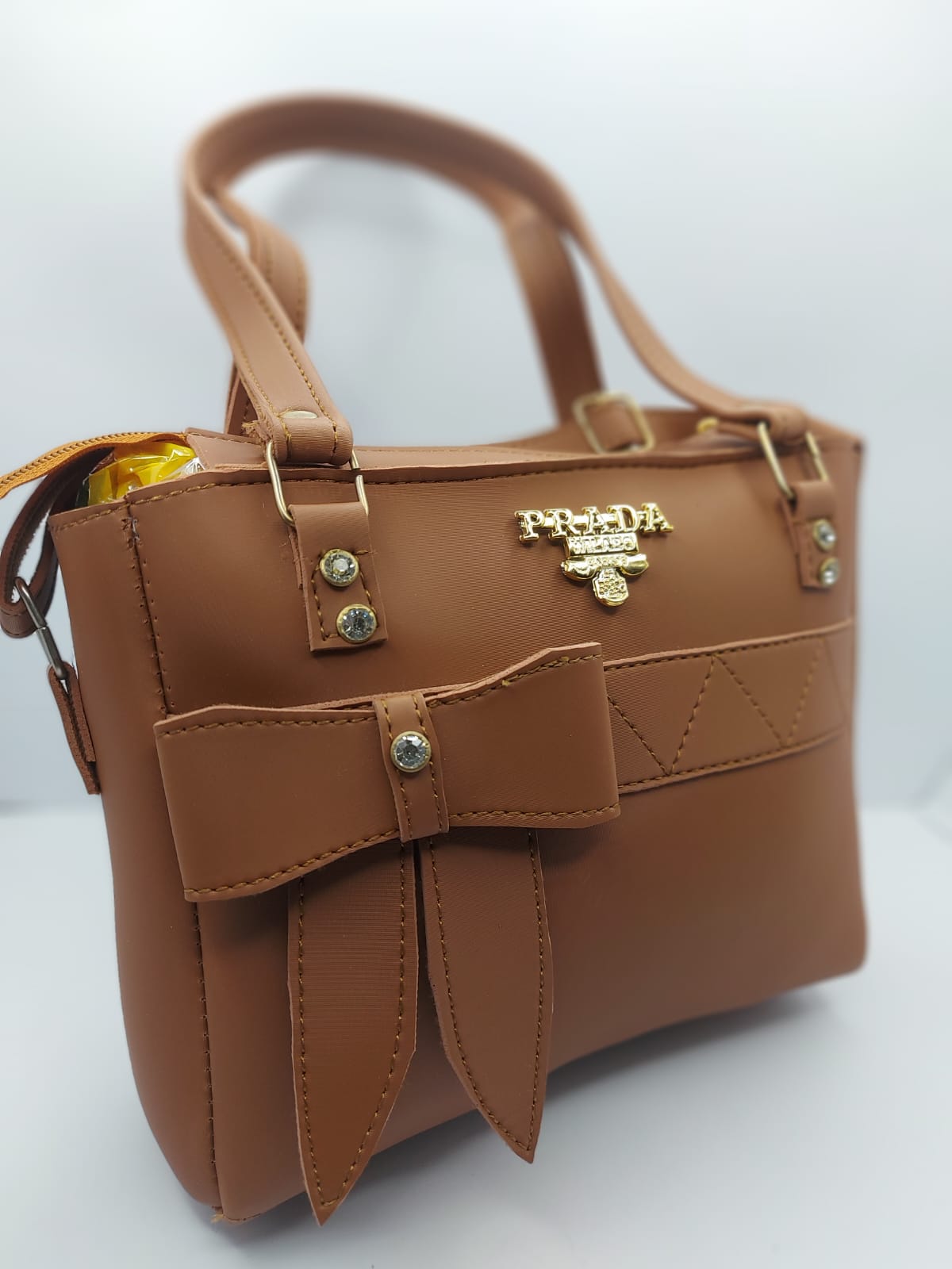 Luxurious camel brown tote bag with a silk-textured finish, bow charm accent, gold-tone Prada-inspired logo, and dual carrying options for versatile styling