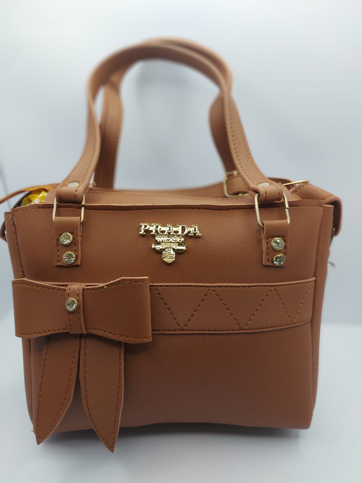 Luxurious camel brown tote bag with a silk-textured finish, bow charm accent, gold-tone Prada-inspired logo, and dual carrying options for versatile styling