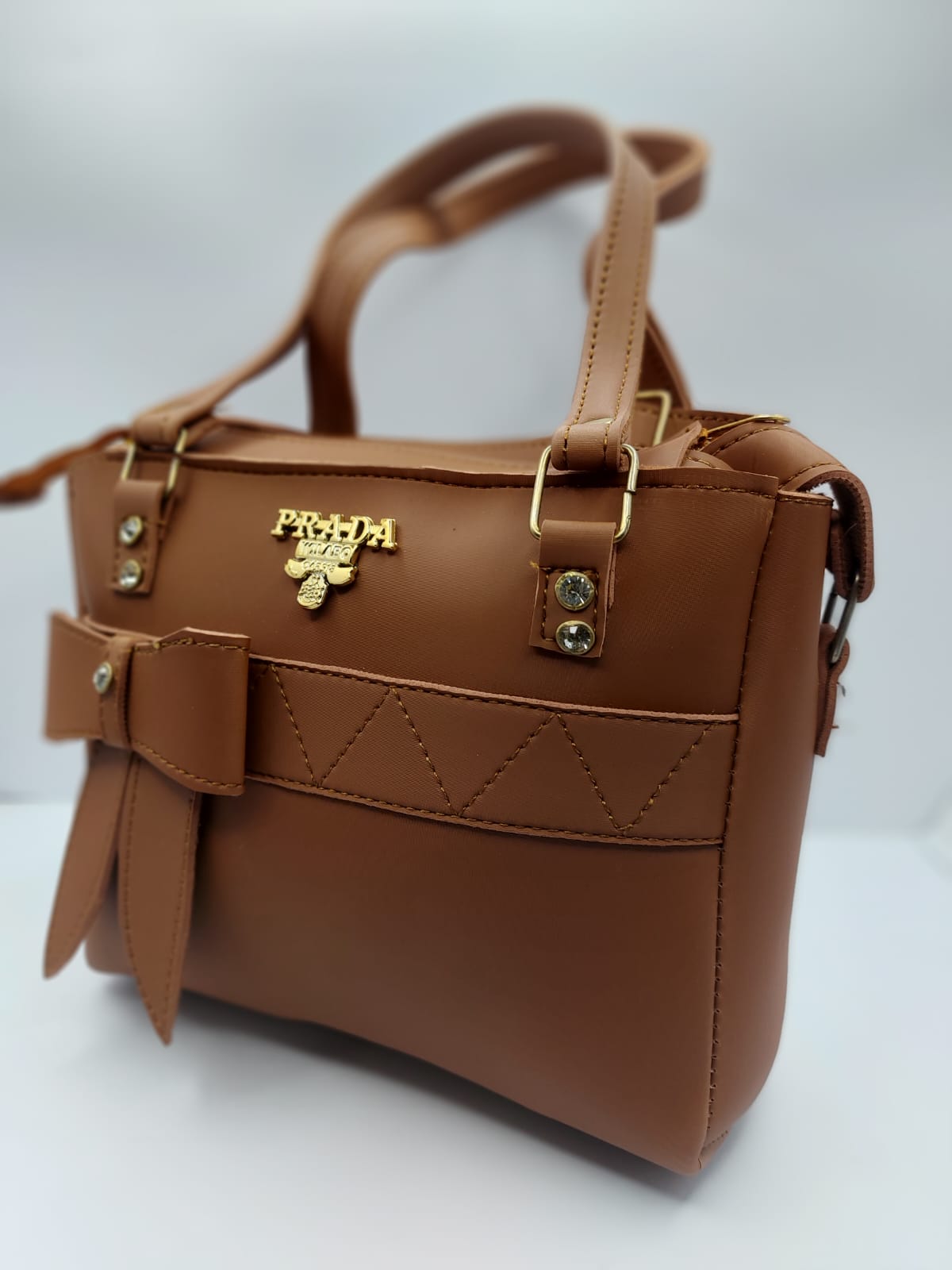 Luxurious camel brown tote bag with a silk-textured finish, bow charm accent, gold-tone Prada-inspired logo, and dual carrying options for versatile styling
