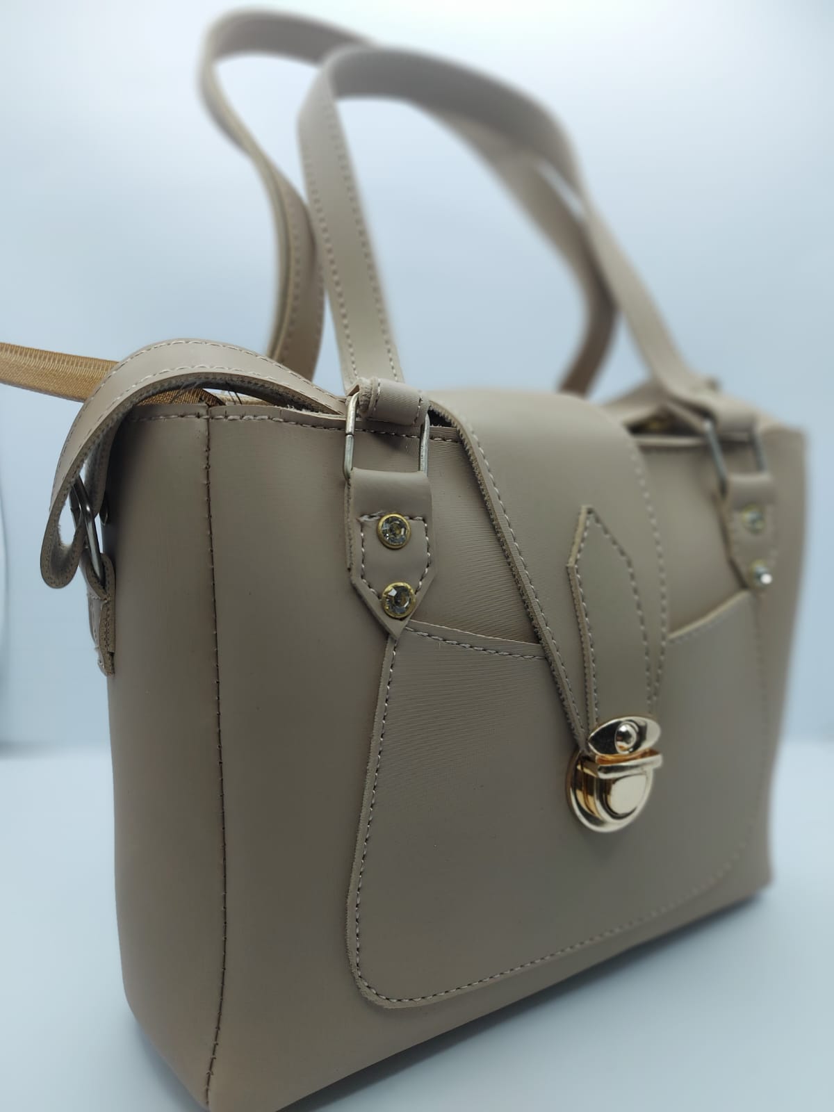 Warm Beige Regal Carry Tote Bag made of premium synthetic leather with neat stitching. Features a gold-tone lock closure, a front pocket for easy access, and an internal zippered compartment for organization. Offers dual carrying options: hand tote and adjustable shoulder strap